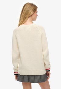 Jersey de punto de color crema con tejido texturizado, que presenta puños acanalados con rayas rojas y azul marino, y un diseño de corte holgado.