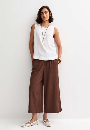 Kobieta ubrana w białą bluzkę bez rękawów, szerokie brązowe spodnie, białe baleriny, długi naszyjnik z zawieszką, bransoletki oraz kolczyki koła, stojąca na tle białej ściany.