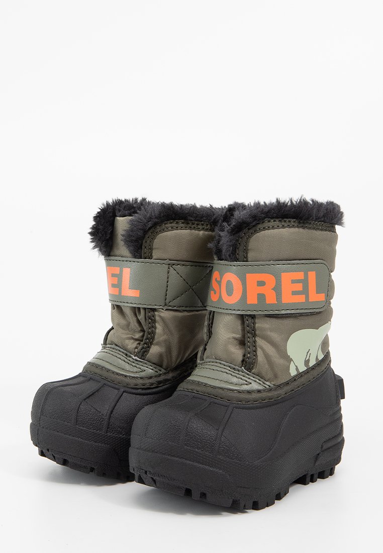 Dunkelgrüne isolierte Stiefel mit schwarzen Gummisohlen, ausgestattet mit einem flauschigen Kragen, orangefarbener "SOREL"-Markierung und einem strukturierten Profil für besseren Halt.