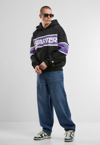 Black hooded sweatshirt with purple and white stripes featuring "STARTER" logo, paired with loose blue denim jeans and green, white sneakers.
