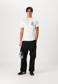 Kurzes weißes T-Shirt mit buntem Grafikdruck, kombiniert mit schwarzen weiten Hosen und schwarzen Sneakers. Hält eine hellblaue Tasche.