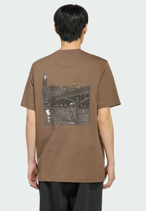 Osoba nosząca brązową koszulkę z grafiką nocnego miejskiego mostu nad wodą, w tym latarni ulicznych i budynków w tle.