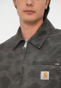 Jakna s dugim rukavima od tamnosive tkanine s uzorkom kamuflaže. Ima zatvaranje na rajsferšlus i mali džep na prsima s Carhartt logom.