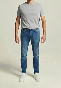 Homme portant un t-shirt gris clair à manches courtes, un jean slim bleu et des baskets blanches, debout devant un fond uni.