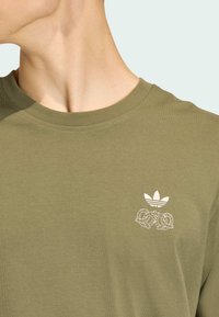 Olivovo zelené bavlnené tričko s okrúhlym výstrihom, s bielym logom Adidas a dvoma prepletenými uzlami na ľavej strane hrudníka.