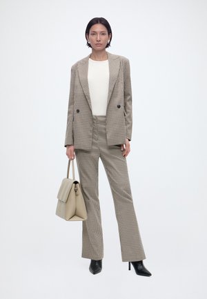 Γυναίκα με μπεζ καρό κοστούμι και λευκό τοπ, κρατώντας μια μπεζ τσάντα και φορώντας μαύρα μπότες με τακούνι, στέκεται μπροστά σε λευκό φόντο.