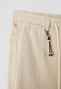 Beżowe lniane spodnie z elastycznym paskiem w talii, wyposażone w sznurek do regulacji oraz dekoracyjną klamrę z koralikami. Tkanina ma delikatny wzór w jodełkę.