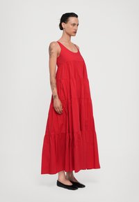 Abito lungo rosso con design senza maniche, volants a strati all'orlo e una texture liscia. Indossato con ballerine nere.