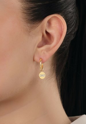 Goldene Creolen mit einem kleinen runden Anhänger, der ein mit Kristallen besetztes Schleifendesign zeigt. Glatte Metalloberfläche, glänzende Verarbeitung.