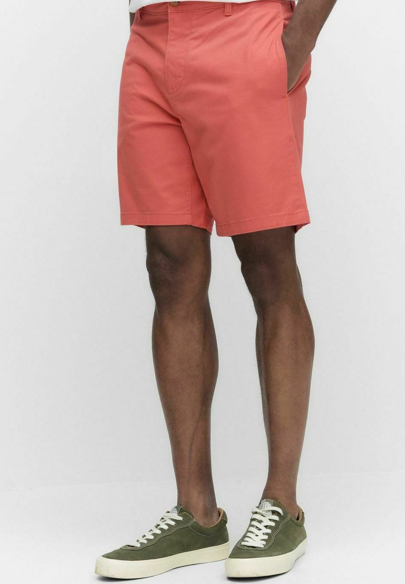 Korallfärgade shorts med rak framsida och sidofickor, tillverkade av lättviktsmaterial. Kombinerade med olivgröna sneakers med vita snören.