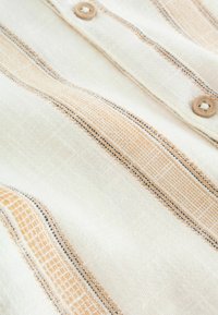 Košeľa z ľahko texturovanej látky s krémovým základom a oranžovo-čiernym pruhovaným vzorom, s drevenými gombíkmi po celej prednej strane.