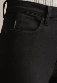 Jean en denim noir avec une texture lisse, un design ajusté, comprenant une poche pour la monnaie, des boutons en métal et des coutures contrastées.