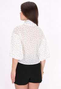 Hvid gennemsigtig bluse med sorte polkaprikker, høj halsudskæring og brede korte ærmer. Sammen med sorte shorts.