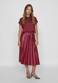 Burgunderfarbene Faltenrock aus Kunstleder, kombiniert mit einem passenden kurzärmeligen Oberteil. Das Outfit zeichnet sich durch eine glatte Textur und einen weichen Fall aus.