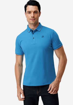 Mann trägt ein hellblaues kurzärmeliges Poloshirt und schwarze Jeans, steht vor einem schlichten weißen Hintergrund.