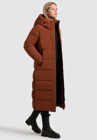 Abrigo acolchado largo marrón con capucha, con un diseño acolchado y cremalleras laterales. Combinado con botas negras hasta el tobillo y un atuendo negro.
