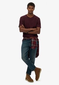 Bordeauxrode T-shirt, blauwe denim jeans en bruine veterlaarzen. Geruit flanellen overhemd om de taille gestrikt. Model staat met gekruiste armen.