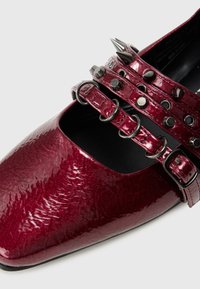 Scarpa in pelle verniciata rossa con punta affusolata, tre cinturini con borchie e spuntoni argentati, e suola nera.