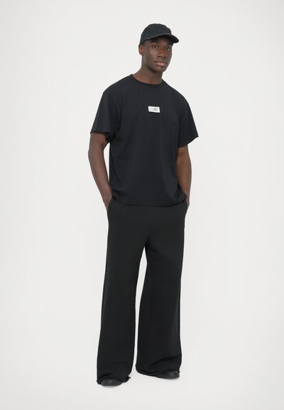 Μαύρο πουκάμισο σε μεγάλο μέγεθος με λευκή λωρίδα λογότυπου, συνδυασμένο με μαύρο παντελόνι φαρδιάς γραμμής. Το ματ ύφασμα και ένα καπέλο ολοκληρώνουν την εμφάνιση.