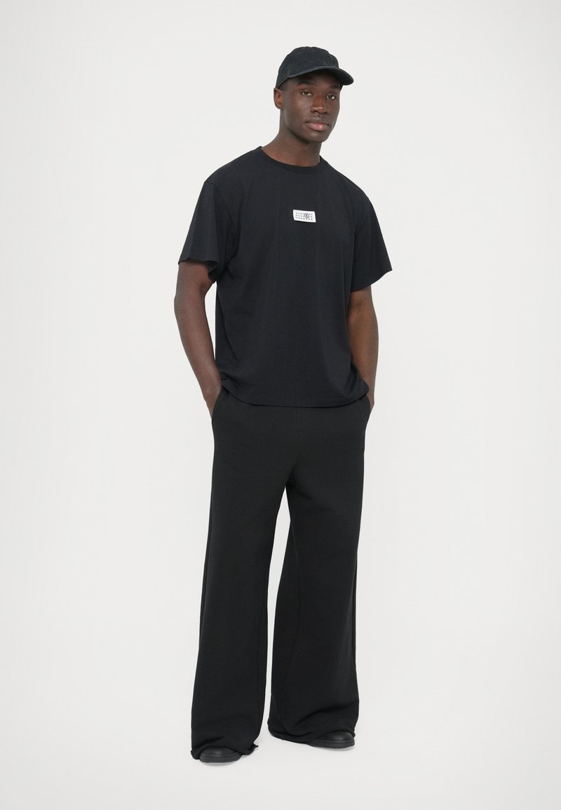 Sort oversized T-shirt med et hvidt logo-patch, kombineret med sorte bukser med vide ben. Matt stof og en kasket fuldender outfittet.
