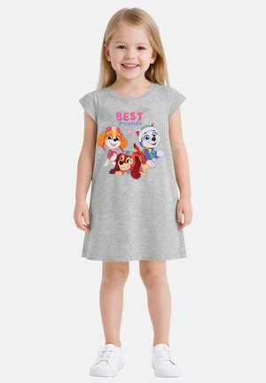 Giovane ragazza sorridente che indossa un vestito grigio con cani dei cartoni animati colorati e la scritta "Migliori Amici", in piedi davanti a uno sfondo semplice.