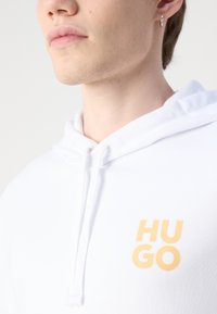 Felpa bianca con cappuccio e cordino, con un logo giallo "HUGO" sul petto. Texture del tessuto liscia, in primo piano sul colletto e sul logo.