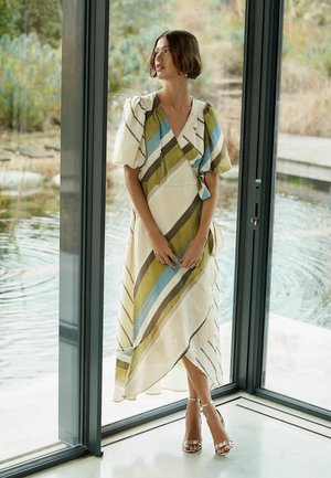 Frau in einem gestreiften Wickelkleid mit Puffärmeln und Knöchelriemen-High Heels, die an Glastüren steht und auf einen Teich und Grünflächen blickt.