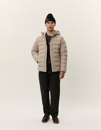 Ung mann står iført beige puffjakke, svart lue, mørkebrun skjorte, svarte bukser og mørkebrun sko mot en ensfarget bakgrunn.
