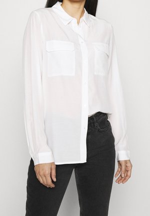 Kvinde iført en hvid skjorte med lange ærmer med forlommer og sorte jeans foran en ensfarvet baggrund.