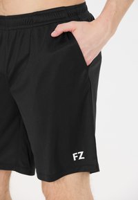 Mies, joka on pukeutunut mustiin urheilushortseihin, joissa on elastinen vyötärö, käsi lepäämään sivutaskussa, "FZ"-logo vasemmassa lahkeessa lähellä saumaa.