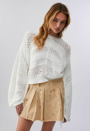 Femeie purtând un pulover tricotat alb cu modele deschise și o fustă plisată bej împodobită cu mici motive florale și stele.