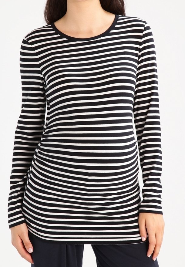 Kvinna i långärmad svart och vit horisontellt randig figurnära tröja och mörka byxor, stående mot en enkel bakgrund.