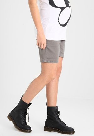 Person iført grå shorts, en hvid t-shirt med sort design og sorte snørestøvler, stående mod en ensfarvet lys baggrund.
