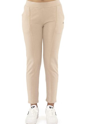Modèle portant un pantalon beige ajusté à taille haute et des baskets blanches, sur un fond blanc uni.