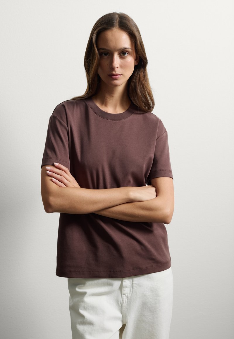 T-shirt in cotone marrone con collo rotondo e maniche corte. Texture liscia, vestibilità rilassata e design minimalista su uno sfondo chiaro.