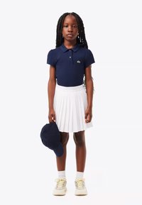 Marinblå polo med kort ärm, vit plisserad kjol, marinblå keps i handen och ljusa sneakers; släta texturer och komplementära färger.