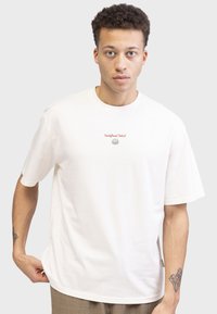 T-shirt blanc en coton à manches courtes, avec un petit logo et un graphique en texte rouge à l'avant. Coupe décontractée avec un col rond.