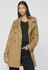 Beige trenchcoat med dubbelknäppt design, slagkragar och svarta knappar, i kombination med en svart topp och byxor. Slät textur.