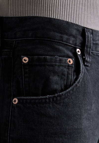 Black denim jeans with a textured surface, featuring a small front pocket with copper rivets and a waistband in gray ribbed fabric.