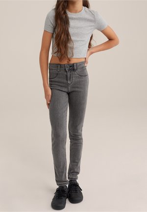 Fille portant un t-shirt court gris, un jean skinny gris et des baskets noires, debout avec une main sur la hanche sur un fond uni.