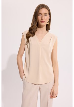 Kobieta o długich brązowych włosach, ubrana w bezrękawową beżową bluzkę z dekoltem w serek i beżowe spodnie, stojąca z jedną ręką w kieszeni na białym tle.