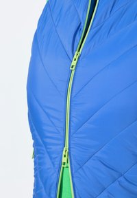 Blå isolerad jacka med diagonalt quiltat mönster och grön dragkedja. Dragkedjan går från halsen till bröstet och avslöjar ett grönt innerfoder.