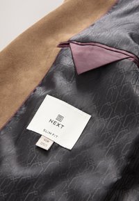 Barna szarvasbőr anyag szürke mintás béléssel; bordó díszítéssel és márka, méret, valamint ápolási információkat tartalmazó címkével.