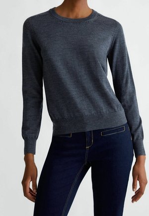Person trägt einen eng anliegenden dunkelgrauen Langarmpullover kombiniert mit dunkelblauen Jeans, steht vor einem schlichten hellen Hintergrund.