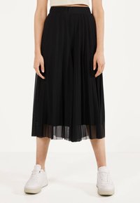 Zwarte geplooide culottes gemaakt van lichtgewicht stof. Heeft een hoge taille, een doorzichtige overlay en een vloeiende silhouet, gecombineerd met witte sneakers.