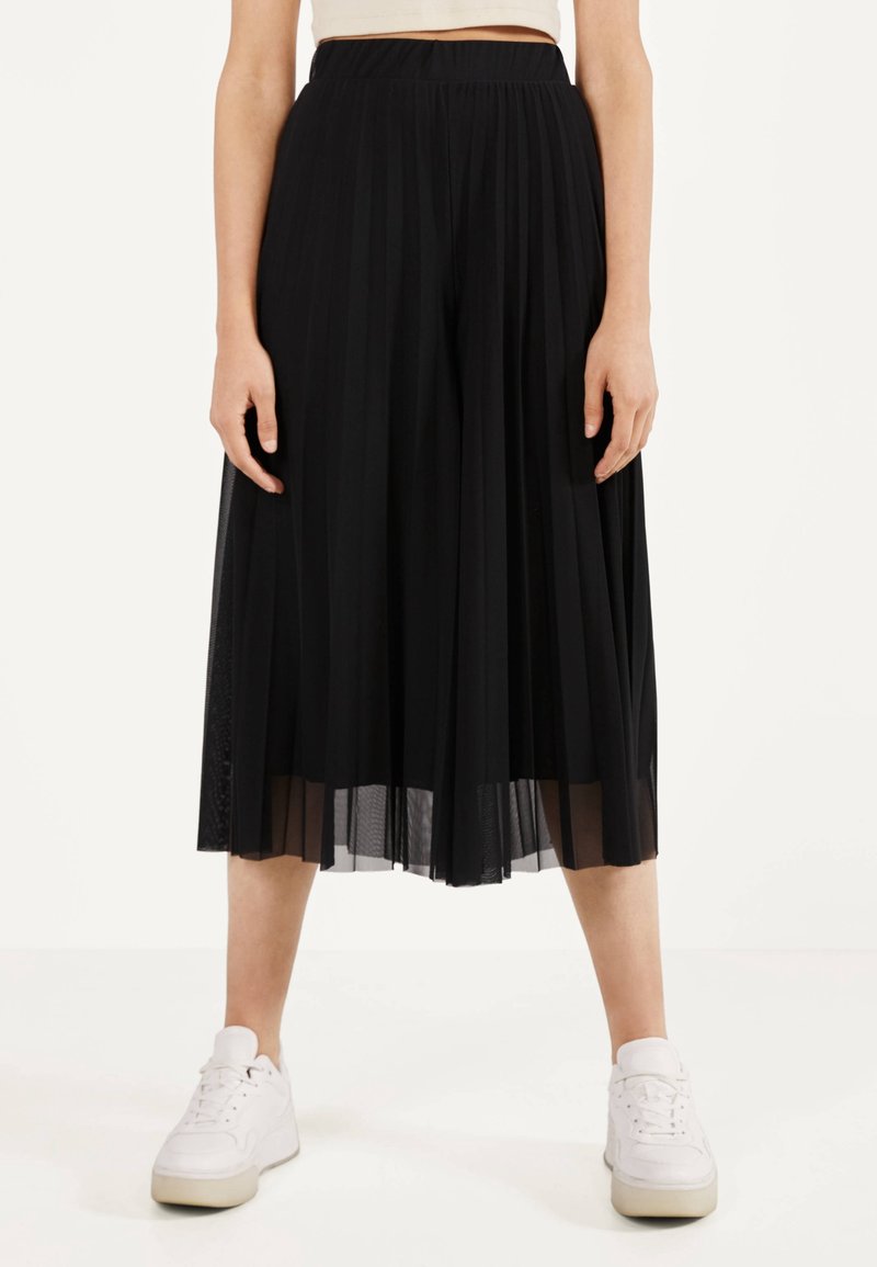 Zwarte geplooide culottes gemaakt van lichtgewicht stof. Heeft een hoge taille, een doorzichtige overlay en een vloeiende silhouet, gecombineerd met witte sneakers.