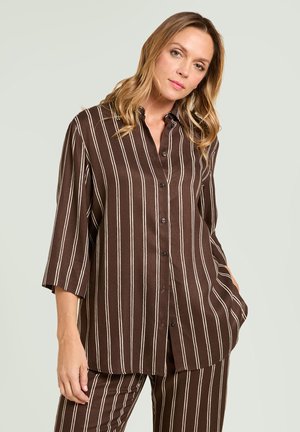 Femme aux cheveux blonds portant une chemise marron boutonnée et un ensemble pantalon avec des rayures blanches verticales, debout devant un fond clair uni.