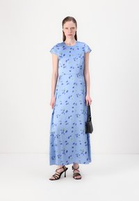 Maxi dress in light blue with floral print. Flared sleeve design, soft fabric, and a straight silhouette. Black handbag and strappy heels.
