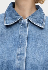 Κοντινό πλάνο ενός ατόμου που φοράει ένα ανοιχτό μπλε τζιν μπουφάν με μυτερό γιακά και ορατές ραφές.