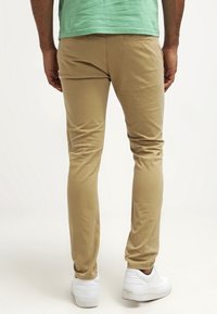 Beige slim-fit chinos vyrobené z bavlny, s kapsami na zadní straně. V páru s bílými teniskami. Pohled ukazuje zadní stranu.